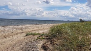 A beach with a pebbly shore, grassy dunes, and a calm sea under a partly cloudy sky. - 6 Person Holiday Home in Roslev (Roslev)