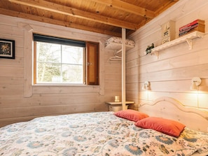 A wooden cabin bedroom with a bed, a window, a shelf with books, and a wall-mounted lamp.