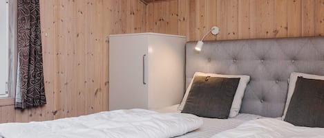 Una habitación con paredes de madera, una cama con ropa blanca, un cabecero gris, una lámpara de noche y un armario blanco.