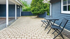 Une terrasse avec un sol en briques, deux chaises en métal noir et une maison bleue avec une fenêtre.