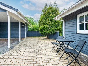 Une terrasse avec un sol en briques, deux chaises en métal noir et une maison bleue avec une fenêtre.