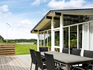 A covered outdoor dining area with a table and chairs, overlooking a grassy field and a body of water.