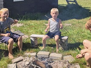 Drei Kinder sitzen um ein Lagerfeuer, im Hintergrund eine hölzerne Konstruktion.