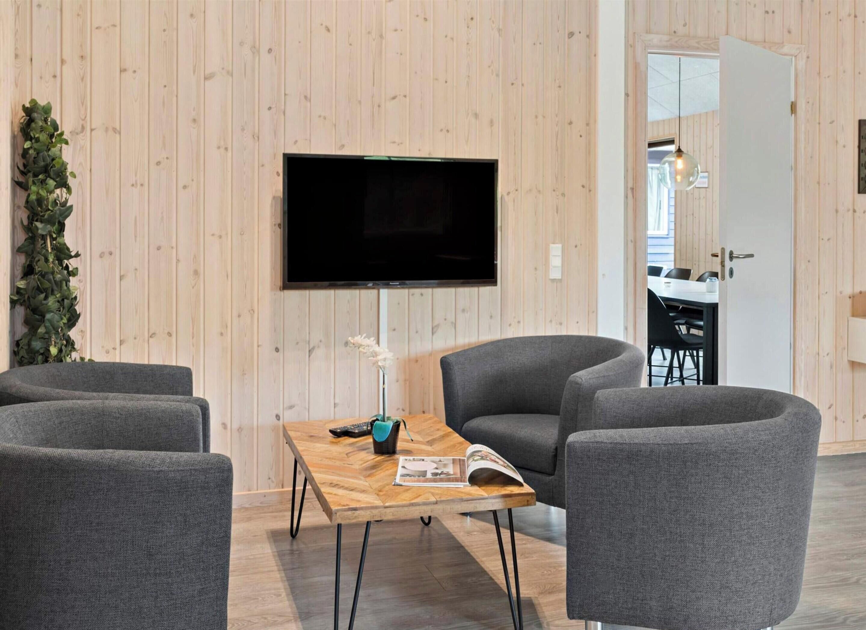 A modern living room with a flat-screen TV, grey armchairs, a wooden coffee table, and a potted plant.