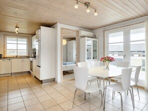 Una cocina moderna con armarios blancos, una zona de comedor con mesa y sillas blancas, y ventanas amplias que dejan entrar luz natural.