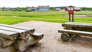 A gravel area with wooden benches and a wooden structure, likely a picnic table, in a rural setting with houses in the background. - 5 Star Holiday Home in Brovst (Brovst)