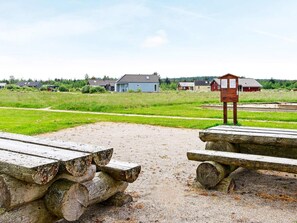 A gravel area with wooden benches and a wooden structure, likely a picnic table, in a rural setting with houses in the background. - 5 Star Holiday Home in Brovst (Brovst)