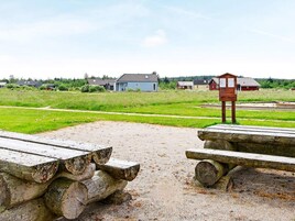A gravel area with wooden benches and a wooden structure, likely a picnic table, in a rural setting with houses in the background.