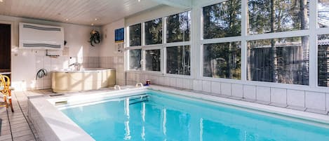A swimming pool with a tiled deck, a built-in hot tub, and large windows offering a view of trees.