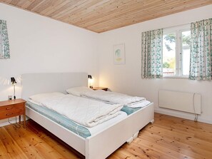 Un dormitorio con techo de madera, una cama con ropa blanca, una mesita de noche con lámpara y una ventana con cortinas florales.