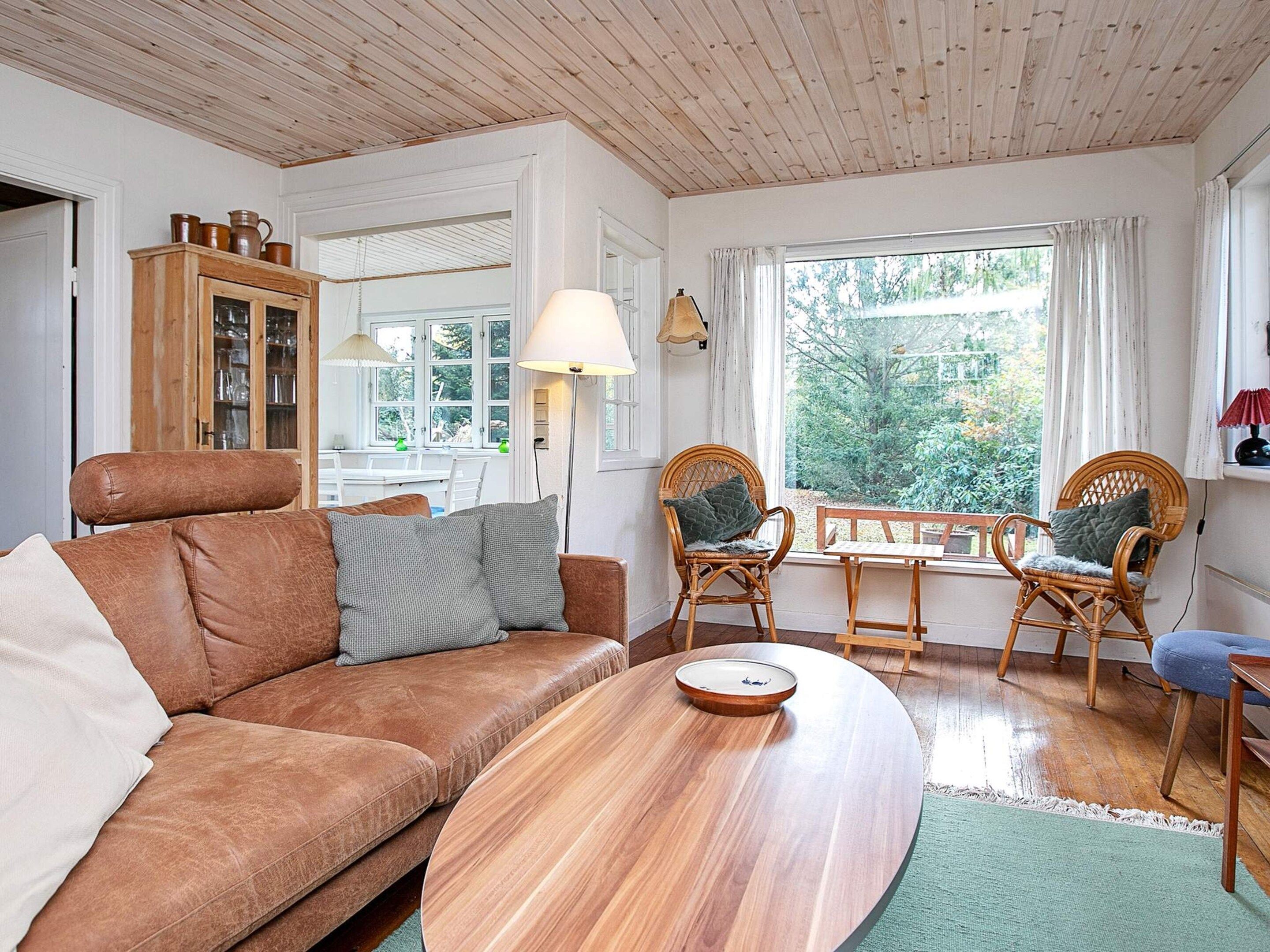 Ein gemütliches Wohnzimmer mit einer braunen Ledersofa, zwei Korbstühlen, einem Holztisch und Blick ins Grüne durch ein Fenster.