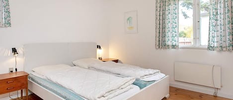 A bedroom with a wooden ceiling, a bed with white bedding, a bedside table with a lamp, and a window with floral curtains.