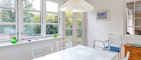 A room with a white table and chairs, large windows, a hanging light fixture, and a wooden cabinet.