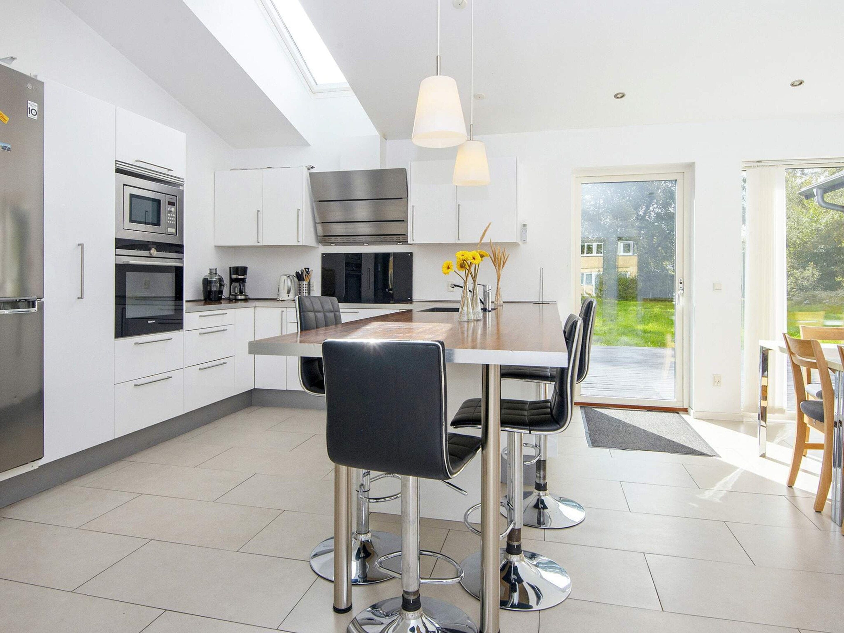 A modern kitchen with stainless steel appliances, a dining area with a large table and chairs, and a view of a garden through a glass door.