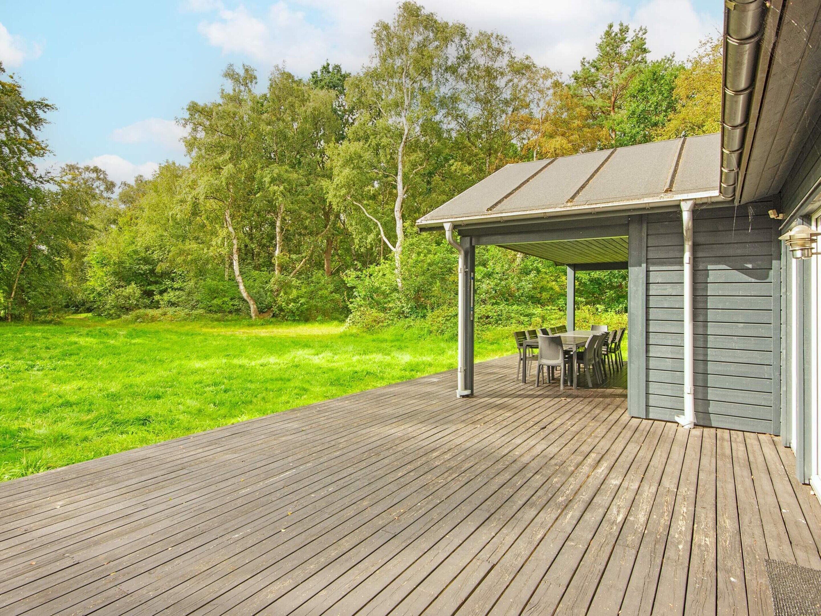 Una terraza de madera con un porche cubierto, mobiliario exterior y vistas a un campo con árboles.