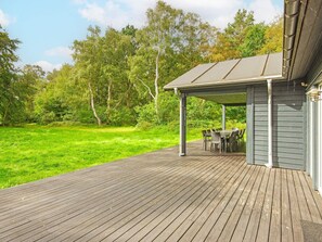 Una terraza de madera con un porche cubierto, mobiliario exterior y vistas a un campo con árboles.
