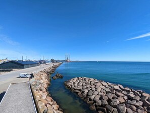 A coastal road with a rocky breakwater, a clear blue sky, and industrial buildings in the background. - 5 Star Holiday Home in Grenaa (Grenaa)
