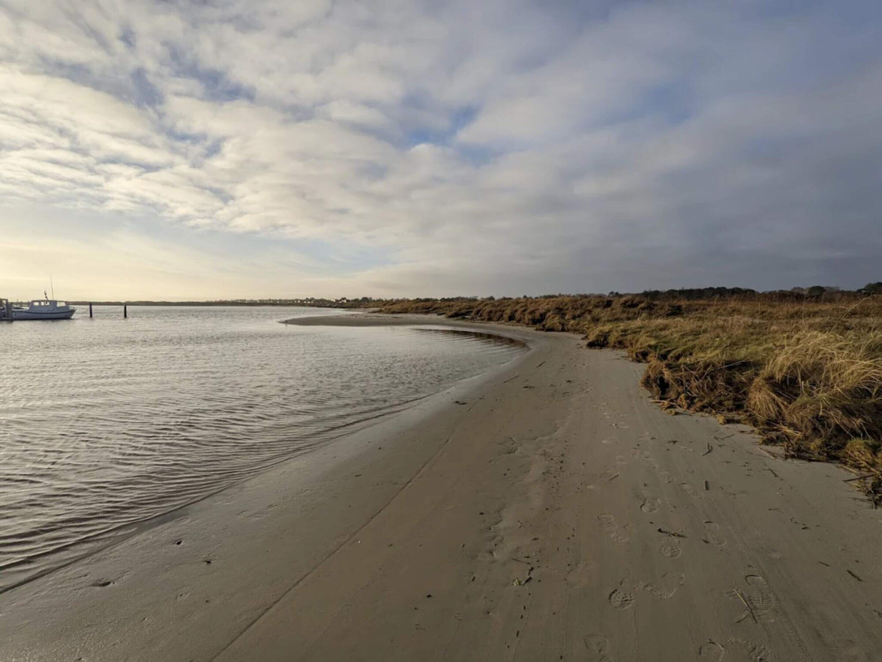 A sandy beach with a boat docked on the left, a curved shoreline, and a grassy dune area.