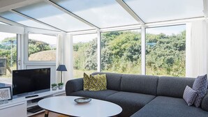 A modern living room with a sectional sofa, a flat-screen TV, a small table, and a large window offering a view of greenery. - 4 Person Holiday Home in Lokken (Lokken)