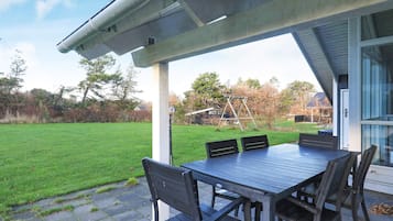 Een terras buiten met een tafel en stoelen, een schommelset en een gazon.