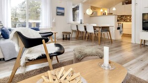 Un salon moderne comprenant un canapé, des fauteuils, une table à manger et un coin cuisine visible à travers une porte ouverte.