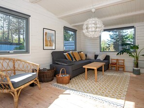 A cozy living room with a sofa, a coffee table, and a wicker chair. There are two windows, a hanging light fixture, and a framed picture on the wall.