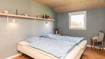 A bedroom with a bed, a window, a chair, and a shelf with books and bottles.