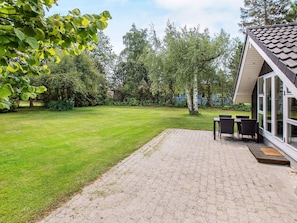 A patio area with a brick floor, a table with chairs, and a view of a garden with trees and a building in the background. - 4 Star Holiday Home in Vaeggerlose (Vaeggerlose)