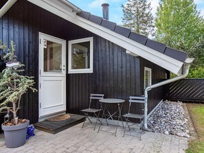 A black wooden building with a white door, two windows, a small outdoor seating area with metal chairs, and a potted plant. - 4 Star Holiday Home in Vaeggerlose (Vaeggerlose)