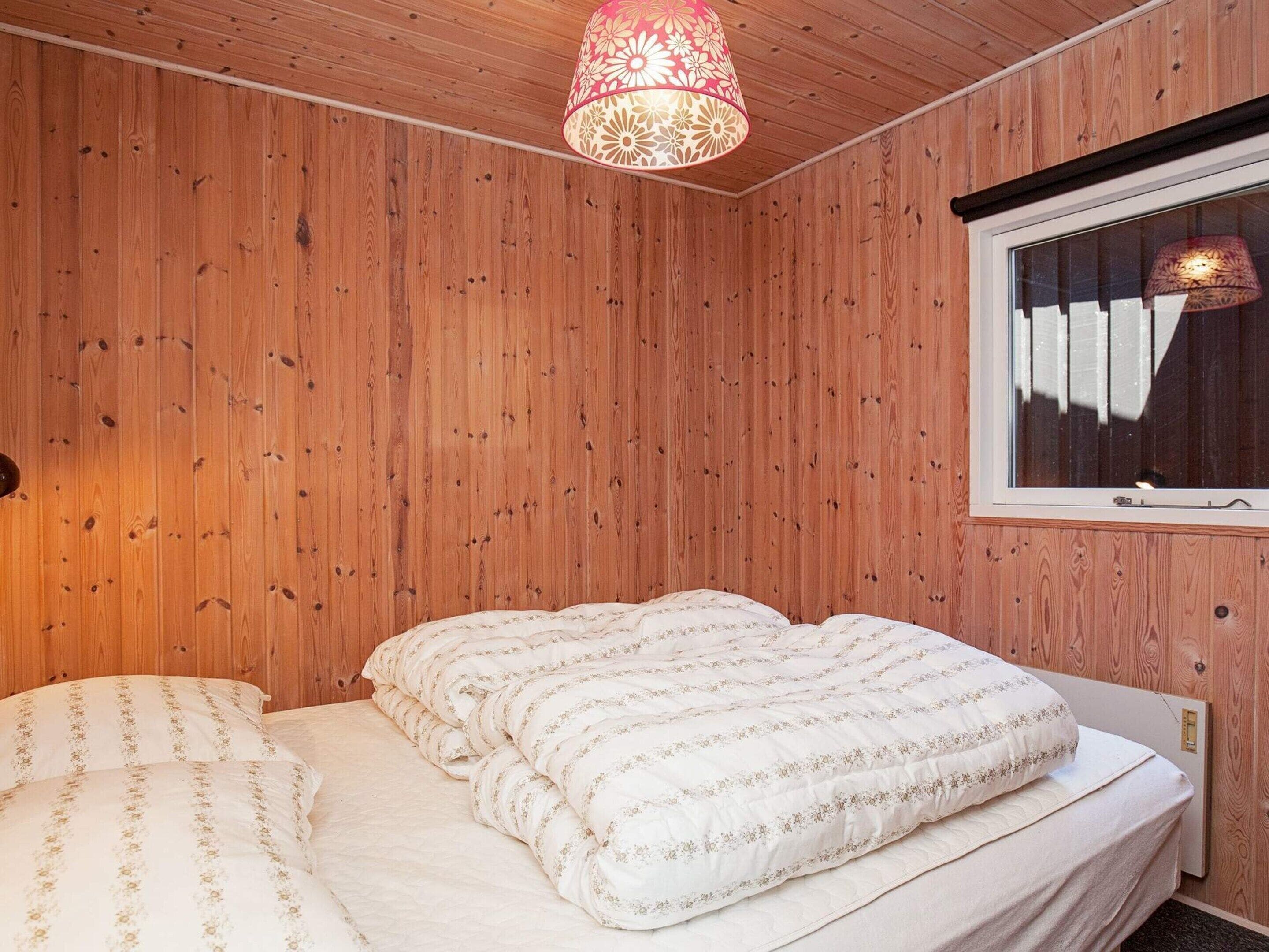 Une chambre avec des murs en bois, un lit recouvert d’une literie à motifs floraux, une fenêtre avec des stores et une lampe suspendue.
