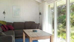 A cozy living room with a wooden table, a brown sofa, and a large window offering a view of greenery. - Spacious Holiday Home in Oksbol near Sea (Oksbol)