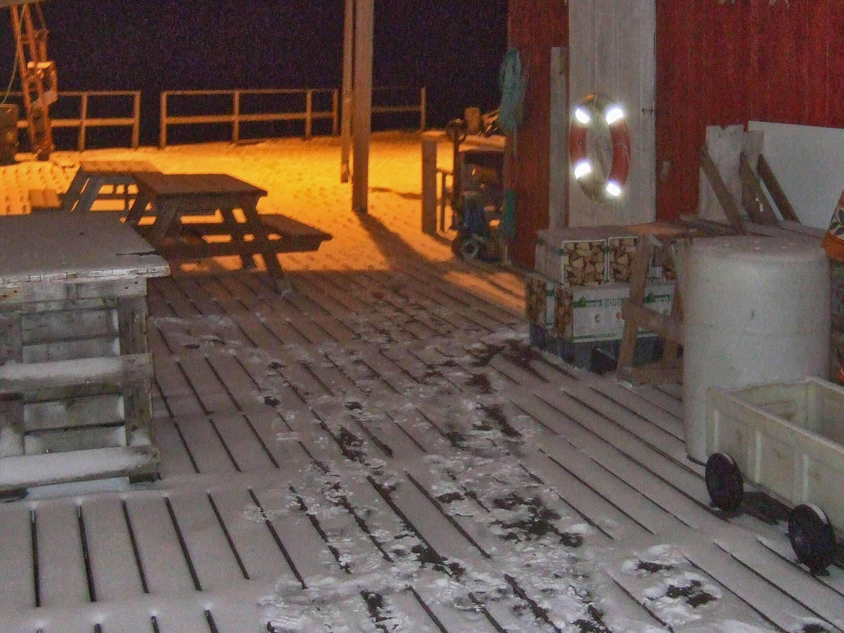 Une terrasse en bois recouverte de neige, une table de pique-nique et un conteneur blanc.