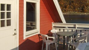 A red building with a white door and window, a white railing, and a table with chairs on a deck overlooking a body of water and a forested hillside. - 4 Person Holiday Home in Rebbenes (Tromsø)