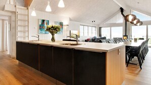 A modern kitchen with a large island, pendant lighting, and a view of the living area with a fireplace. - 5 Star Holiday Home in Ebeltoft (Ebeltoft)
