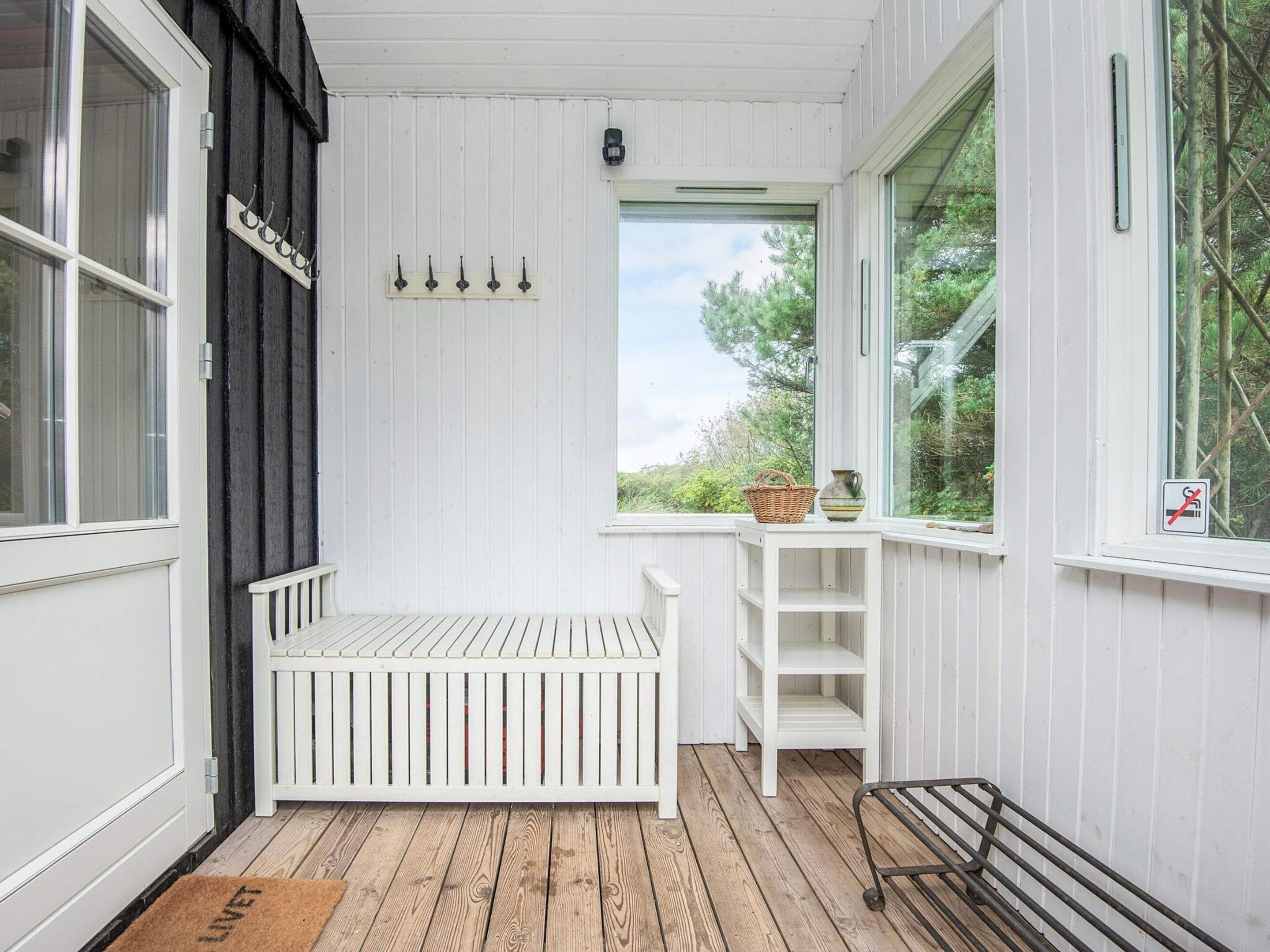 A white wooden room with a bench, a window with a view of greenery, and a door with a sign.