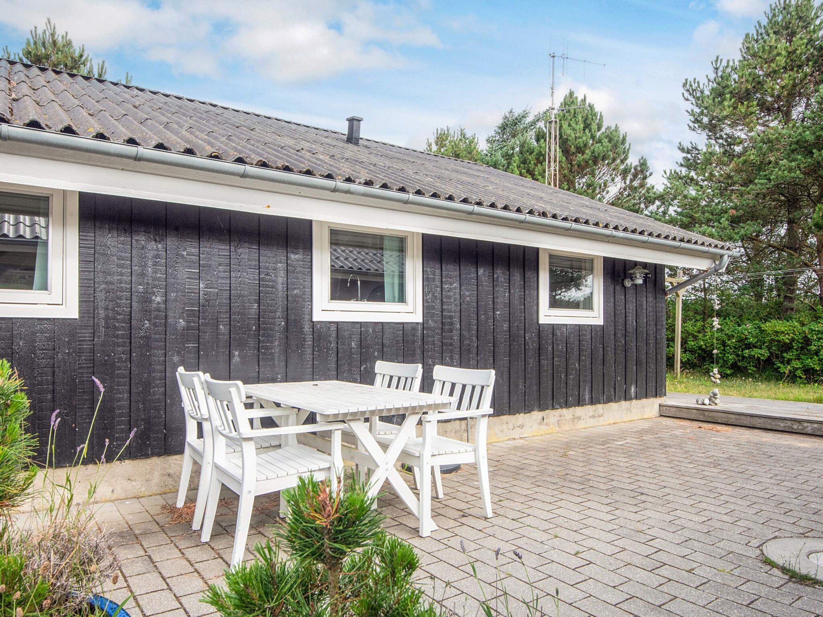 A small house with a dark exterior, white window frames, and a tiled patio with a white table and chairs.