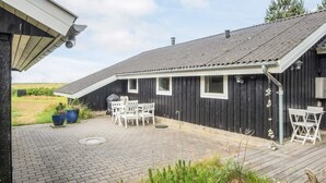 A black wooden building with a sloped roof, a patio with a table and chairs, and a view of the surrounding landscape.