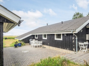 A black wooden building with a sloped roof, a patio with a table and chairs, and a view of the surrounding landscape.