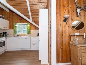 Eine Küche mit weißen Schränken, einem Fenster mit Vorhängen, einer Holzbalkendecke und einer Holzwand mit Spiegel und Wandleuchten.