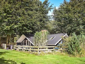 A rustic cabin with a sloped roof, surrounded by dense trees and a wooden fence.