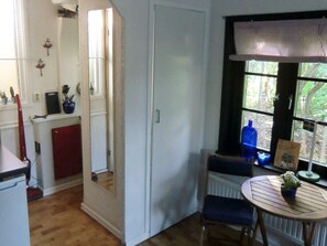 Una habitación con piso de madera, una puerta blanca, una ventana con vista a follaje y una pequeña mesa redonda con una planta, junto a una silla.