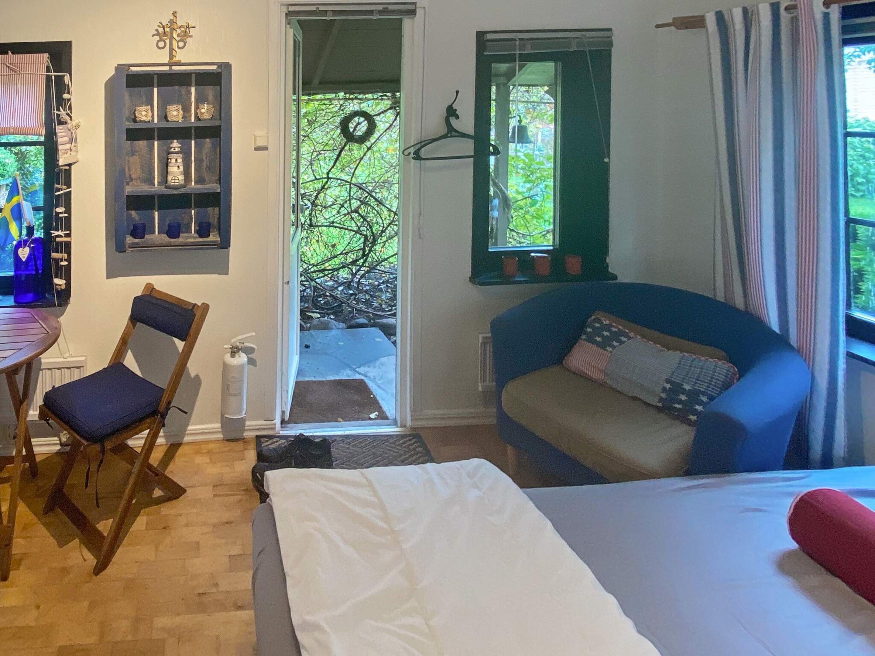 A room with a bed, a blue sofa, a wooden chair, a small table, a shelf with decorative items, and a door leading to an outdoor area.