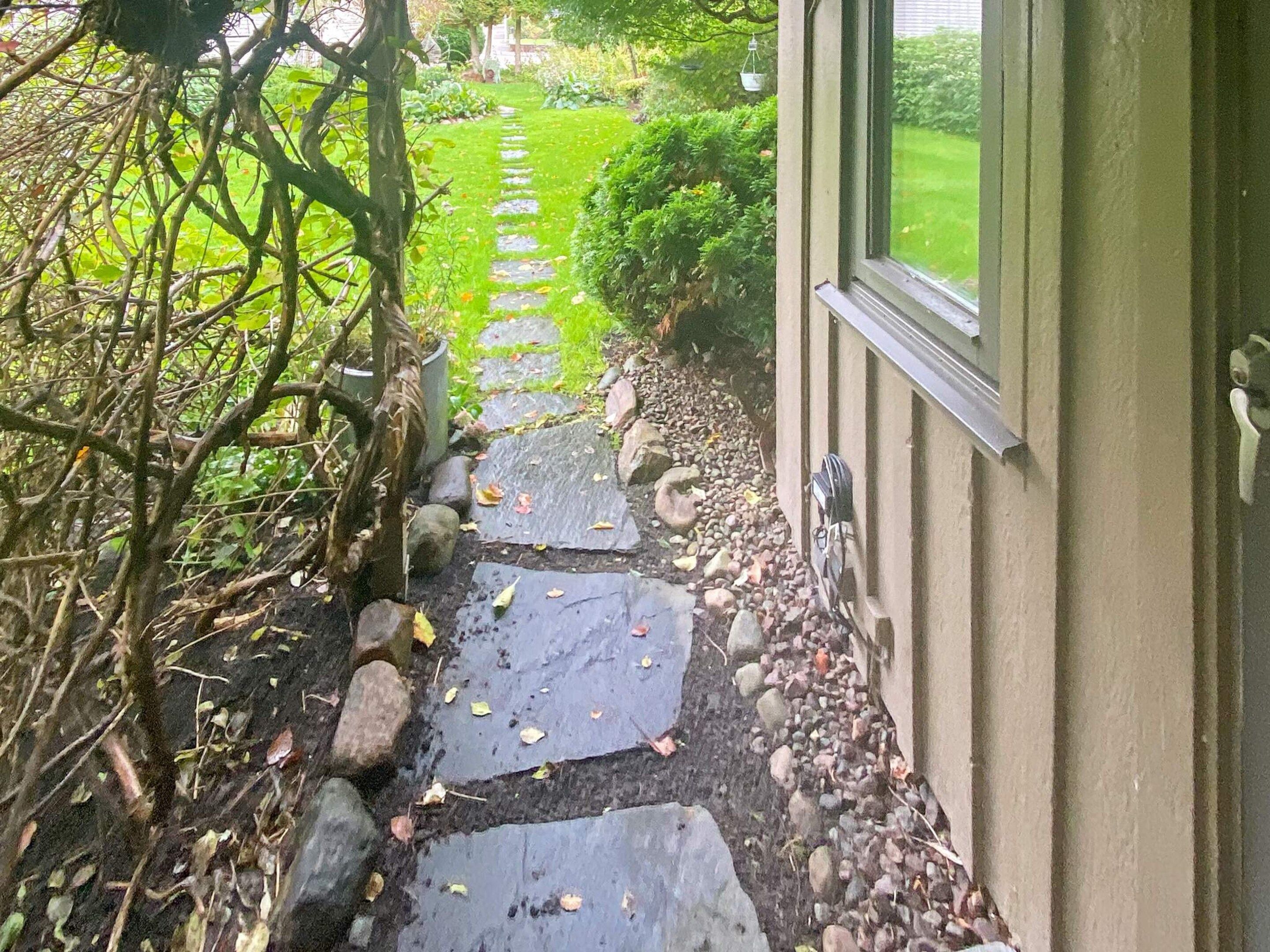 A stone pathway leading to a house with a door and window, surrounded by greenery and a lawn.
