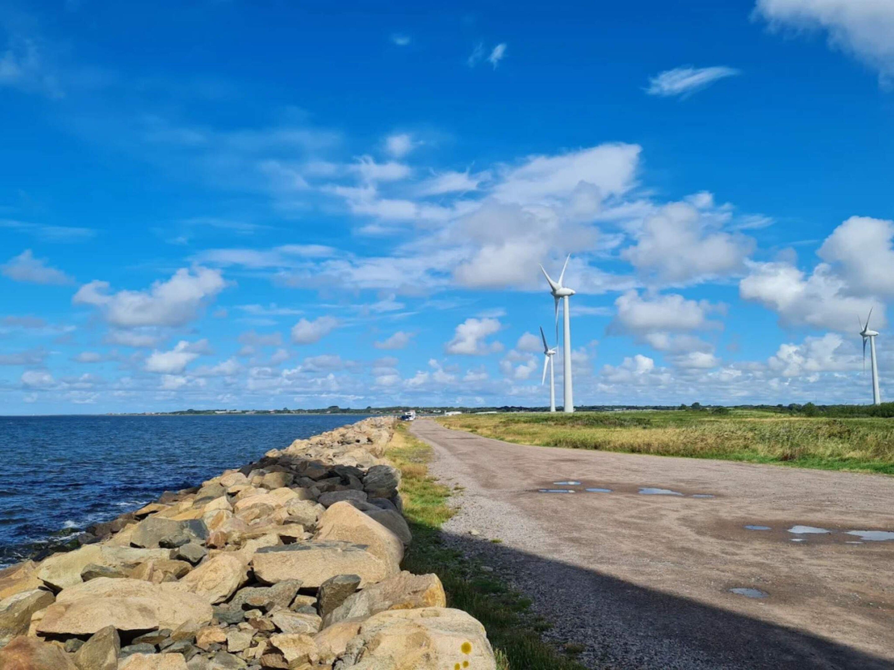 A coastal road with wind turbines in the distance, a rocky shoreline, and a clear blue sky.