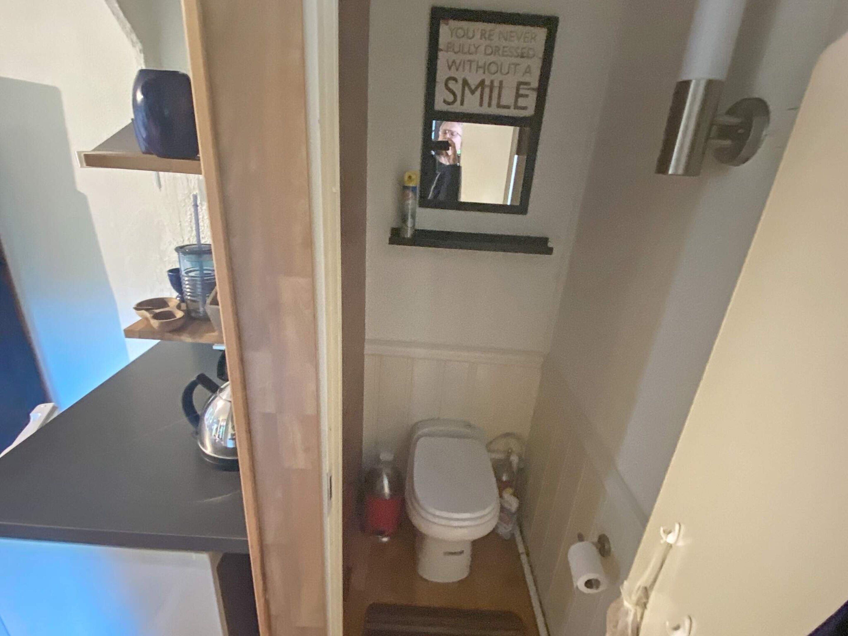 A small bathroom with a toilet, a sink, and a mirror. There is a sign on the wall that reads 'YOU'RE NEVER BULLY DRESSED WITHOUT A SMILE.'