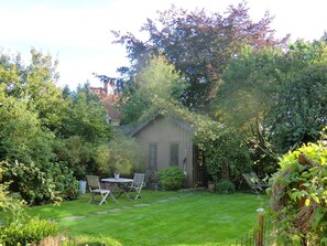 En have med et lille skur, et bord med stole og mange forskellige planter.
