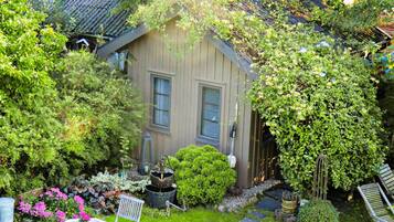 Ett trähus med en trädgård, en liten uteplats med ett bord och stolar samt ett stort utbud av växter och blommor.
