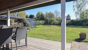 A covered outdoor area with a wooden deck, wicker furniture, and a view of a backyard with a blue shed and greenery. - 4 Star Holiday Home in Dannemare (Dannemare)