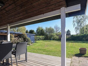 A covered outdoor area with a wooden deck, wicker furniture, and a view of a backyard with a blue shed and greenery.