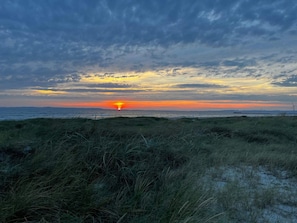 Un coucher de soleil sur une plage, avec des dunes herbeuses et des éoliennes à l’horizon.